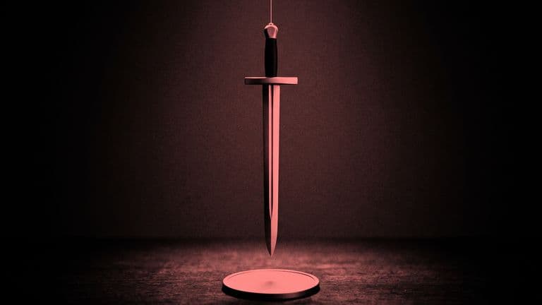 A sword hangs point down on a thin thread just above a blank coin lying on a dark table.