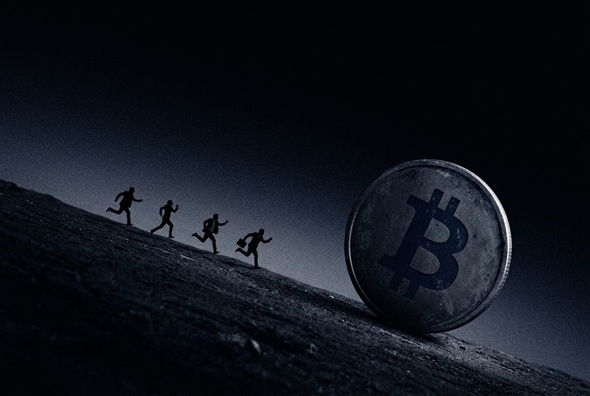 A giant Bitcoin coin rolls down a dark slope while a few small suited figures run downhill after it.