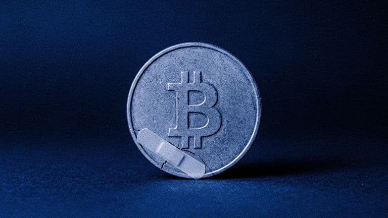 A coin with the Bitcoin mark has a thin crack across it, covered by a sticking plaster, on a flat blue background.