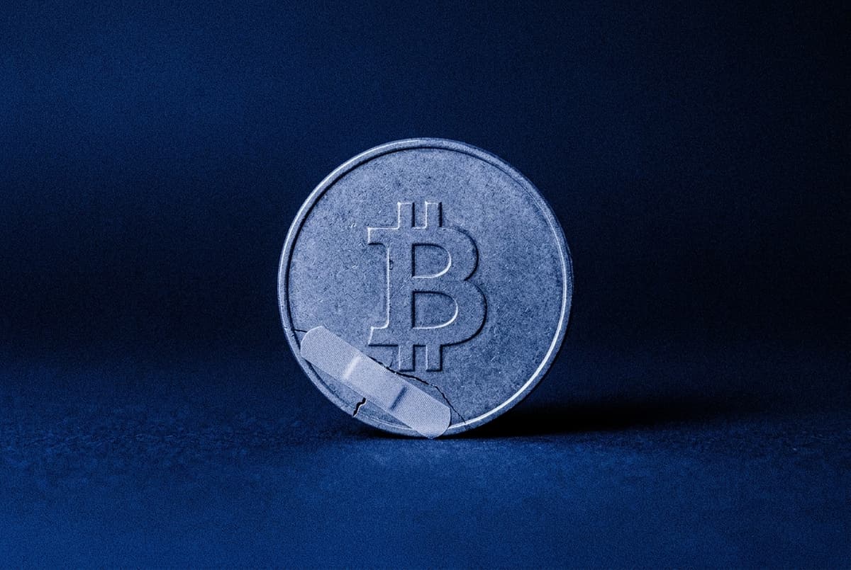 A coin with the Bitcoin mark has a thin crack across it, covered by a sticking plaster, on a flat blue background.