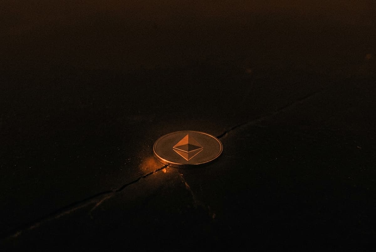 A coin with the Ethereum mark lies on a dark sheet of ice, with a thin crack spreading out from beneath it.