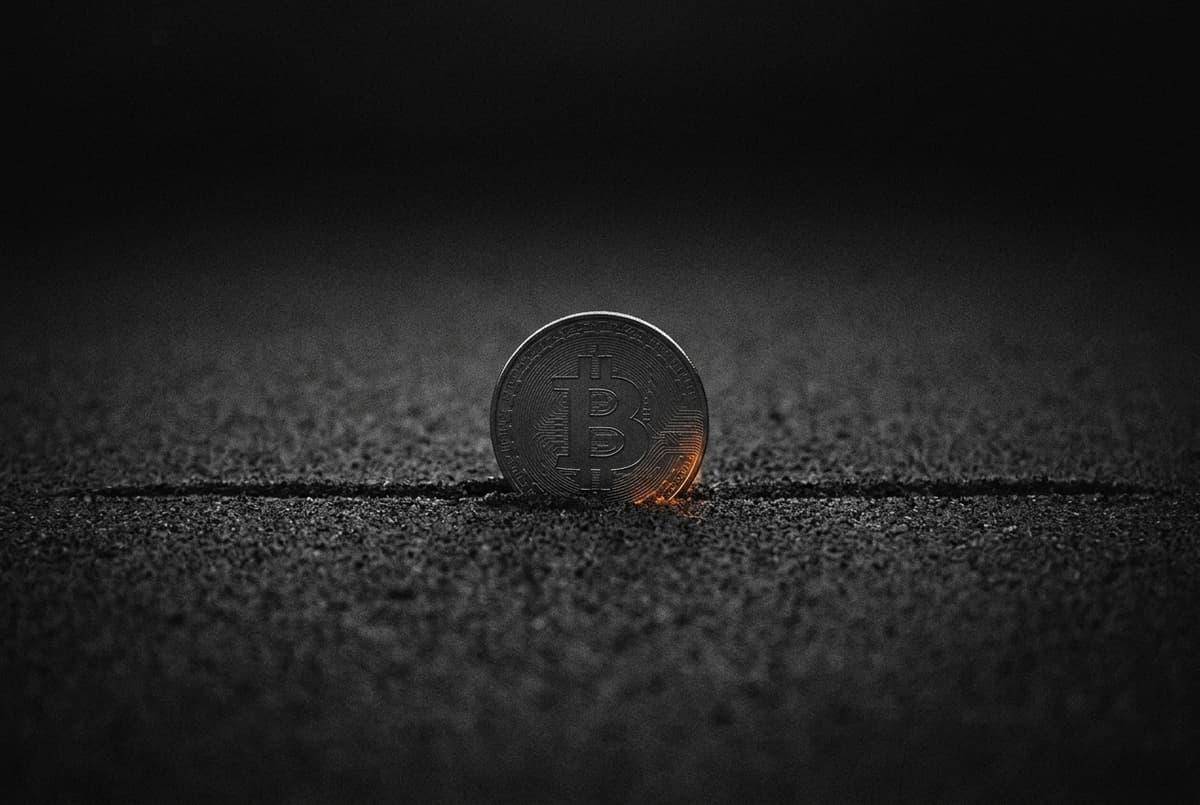 A coin with the Bitcoin mark stands upright in dark sand, resting exactly on a single straight line drawn across the sand, with darkness above.
